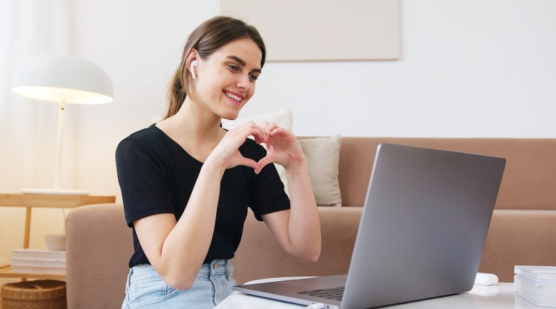 Cheerful happy young female in jeans and black shirt having conversation via video call on laptop and making heart shape with fingers while sitting on floor in modern living room